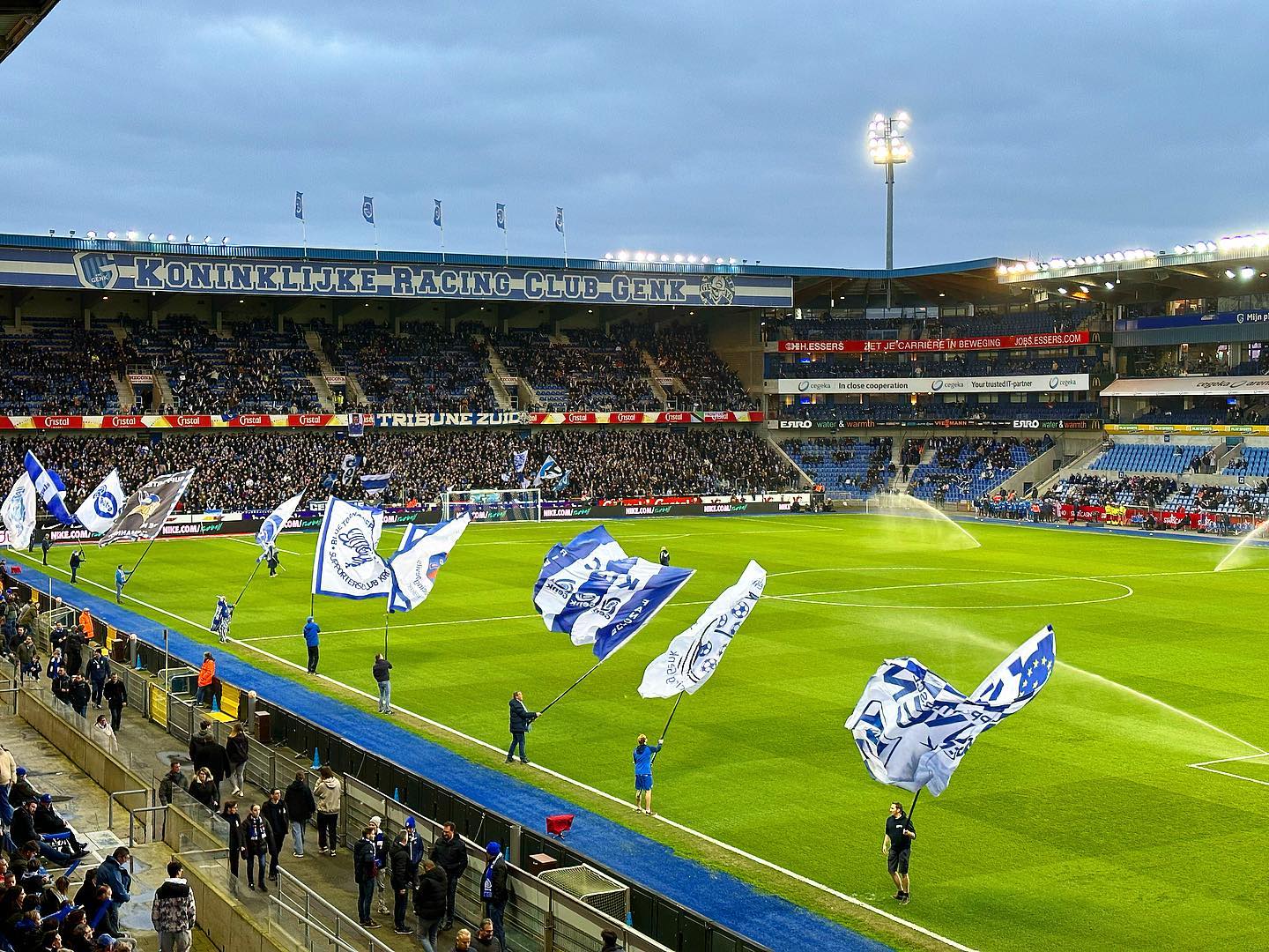 Vol stadion bij KRC Genk met vlaggenzwaaiende supporters en sproeiers op het veld in de Cegeka Arena